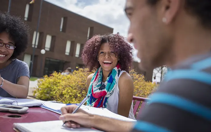 A student smiles at another as they work on a project on a picnic table.