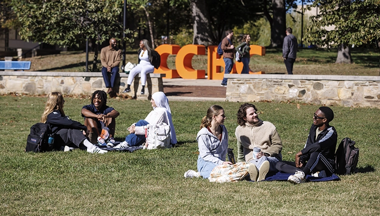 Groups of students are sitting together on a lawn with CCBC letters in the background
