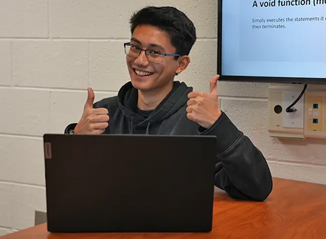 Younger male student sitting behind him laptop giving thumbs up