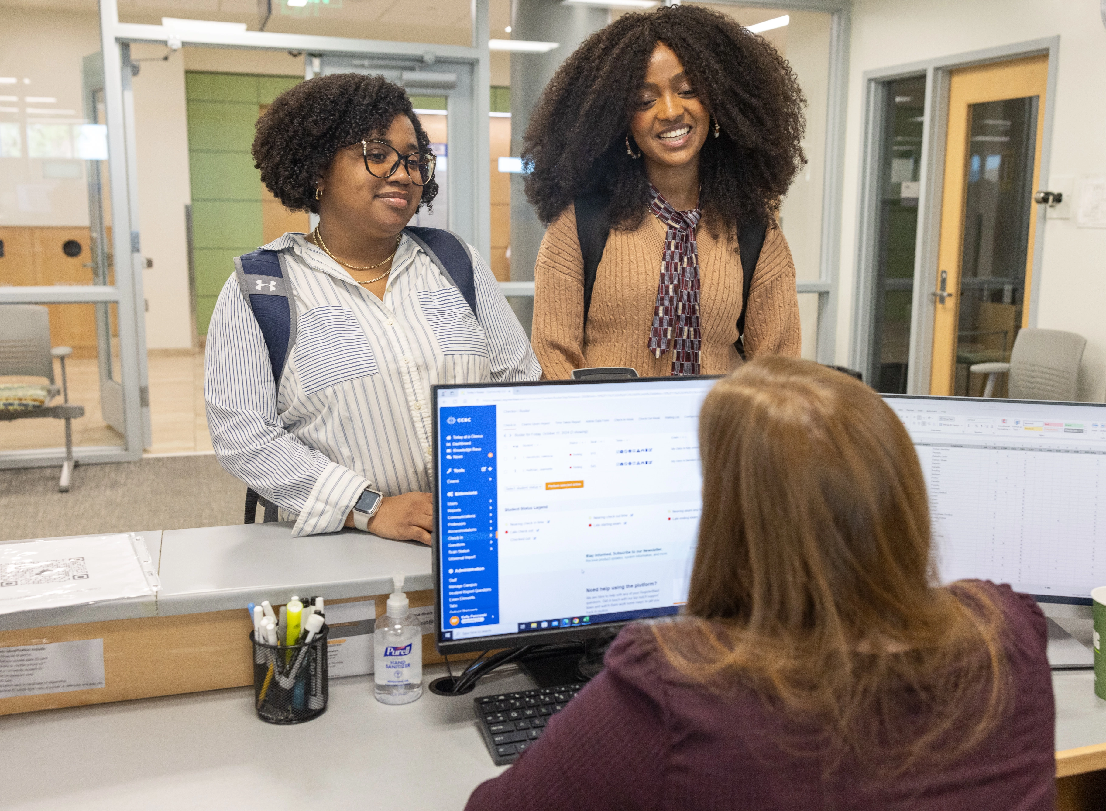 Two students pose in a college hallway to take a picture and smile beneath a sign that reads "Testing Center"