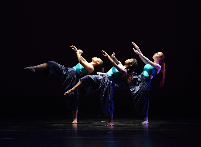 Three dancers against black background dressed in blue