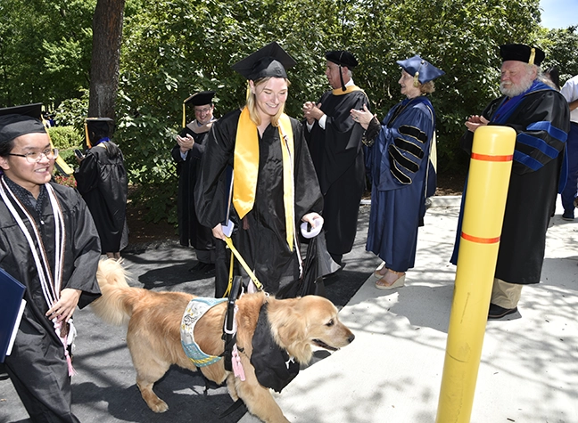 A graduating student dressed in Commencement attire stands in the procession line with their service dog.