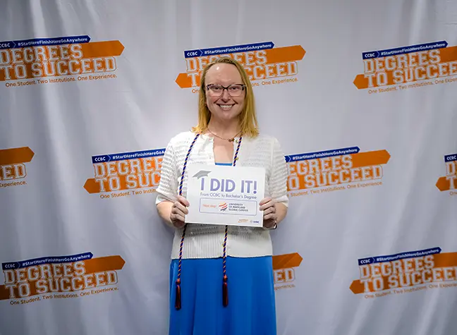A student attends the Degrees to Succeed Commencement Ceremony and holds a sign that reads "I did it!" next to the UMGC logo.