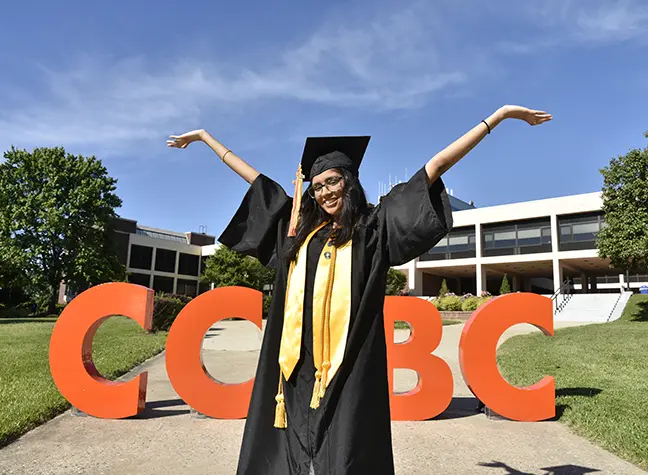 Vidhi Patel stand in front of orange letters that read "CCBC" in her graduation robe.