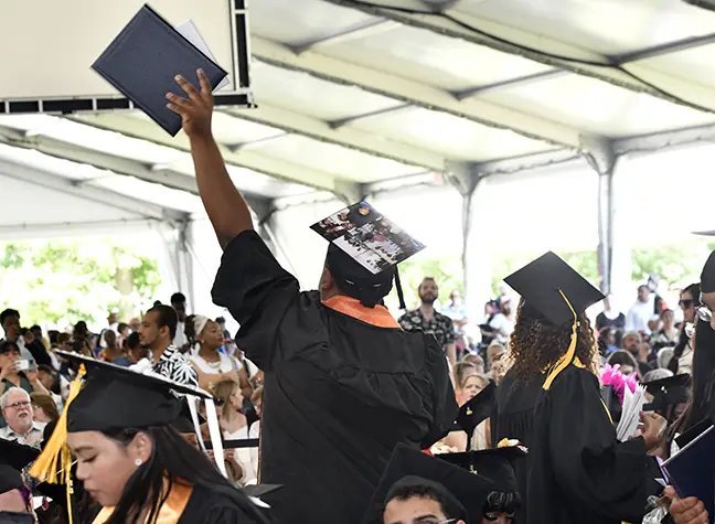 Student in graduation garb with his back to the camera holds degree in the air.